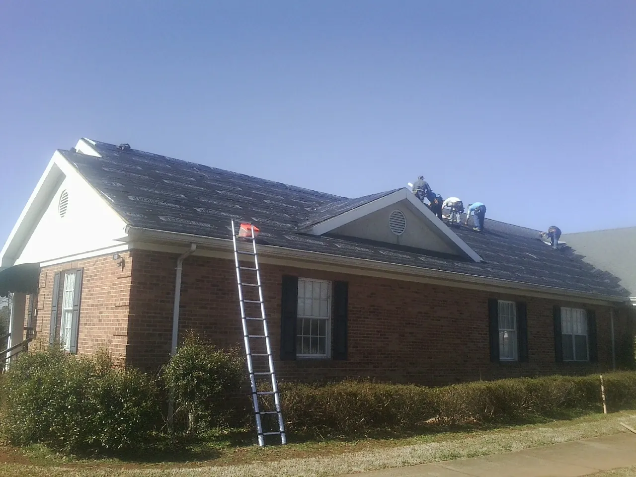 Crew tearing shingles off a brick ranch roof with a ladder set — Metal Roof Repair work by our crew in Cedar Crest