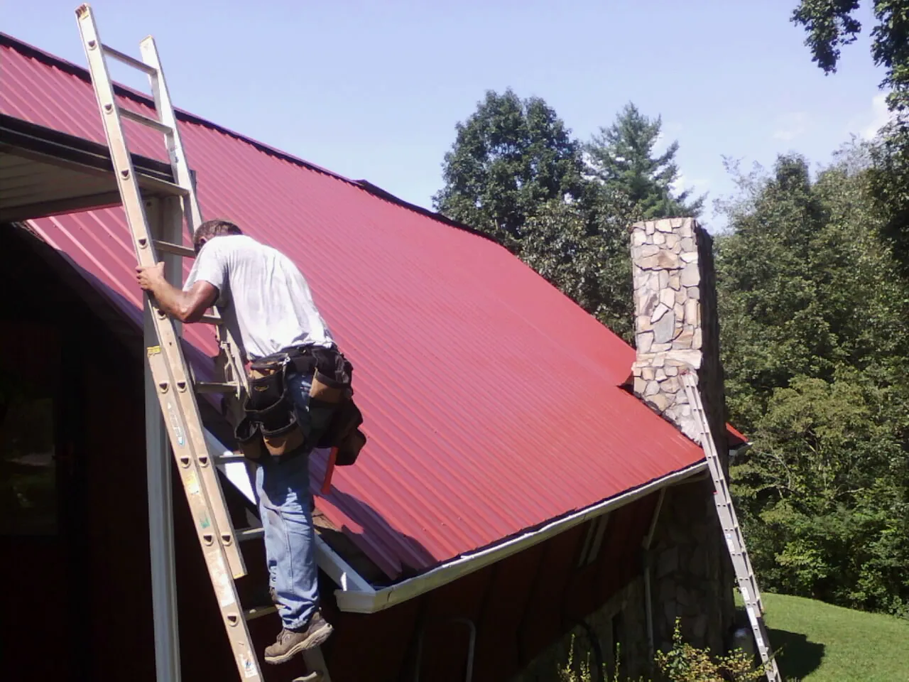 Roofer on a ladder beside a red metal roof and stone chimney — Soffit & Fascia Repair by Roofing Brothers in Cedar Crest